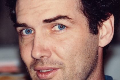 "Man with blue eyes and dark curly hair, wearing a blue shirt, looking directly at the camera in an indoor setting with a blurred background."