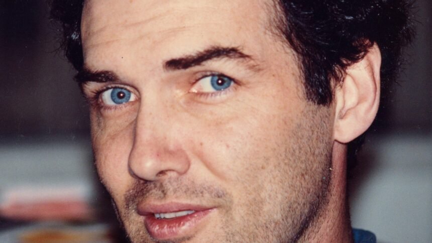"Man with blue eyes and dark curly hair, wearing a blue shirt, looking directly at the camera in an indoor setting with a blurred background."